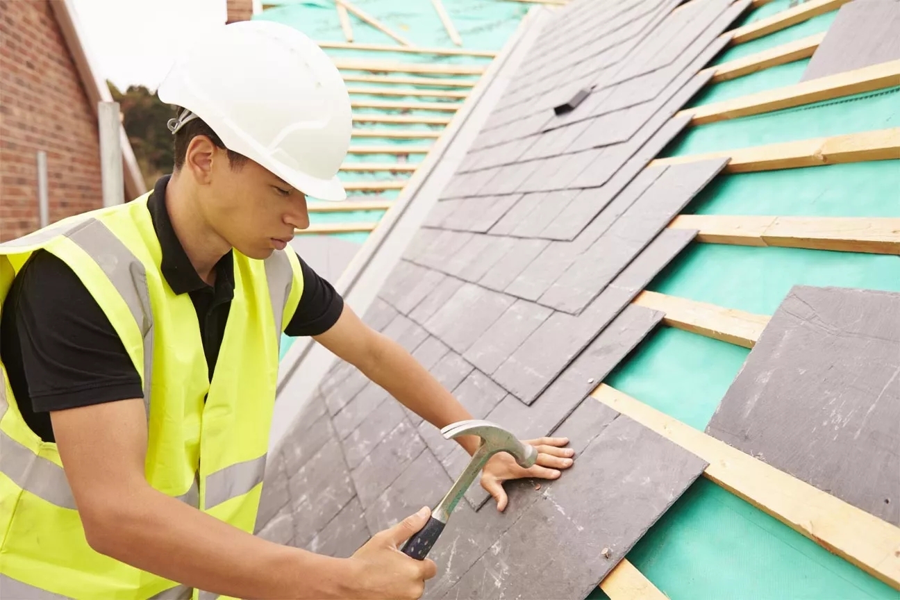 A construction worker wearing a safety jacket and helmet hitting a nail into roof.