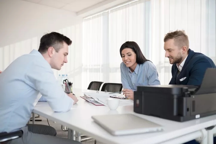 Three people in a meeting at a table discussing an open notebook on the meeting table between them.