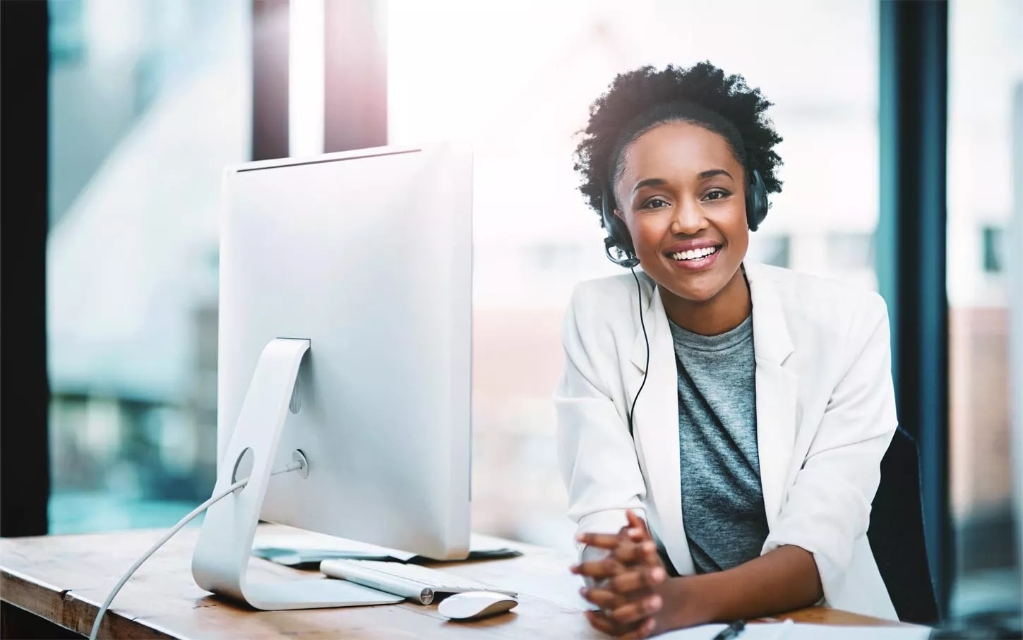 A women wearing a headset and white blazer and has a iMac before her on the table.