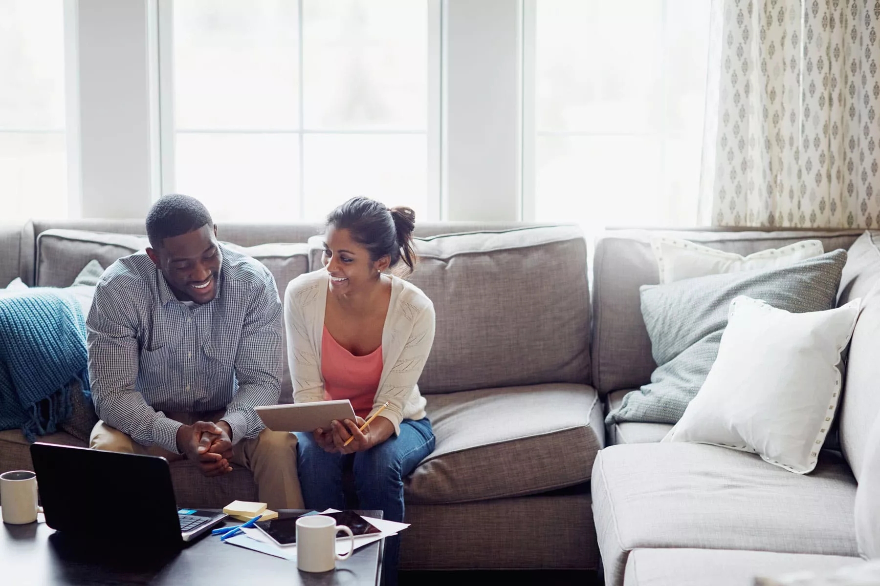 A man and a women with a book and pencil in the hand sitting on a sofa and has laptop before them on a coffee table.